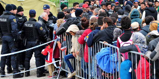 Dutzende Menschen warten an einem Zaun, dahinter Polizisten und ein Zelt. Dutzende Menschen warten an einem Zaun, dahinter Polizisten und ein Zelt.