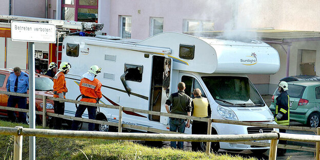 Feuerwehrleute und Polizisten vor dem qualmenden Wohnwagen des NSU