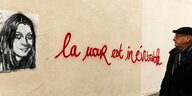 Ein Frauenproträt eines Paris-Opfers an einr Hauswand, daneben ein rotes Grafitti „l'amour est inevitable“ und ein Mann, der es betrachtet