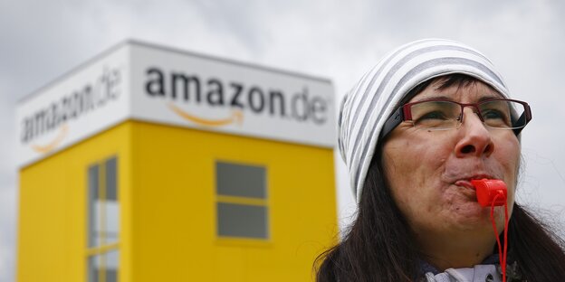 Eine Frau hat eine rote Trillerpfeife im Mund, hinter ihr ein Amazon-Logo Eine Frau hat eine rote Trillerpfeife im Mund, hinter ihr ein Amazon-Logo