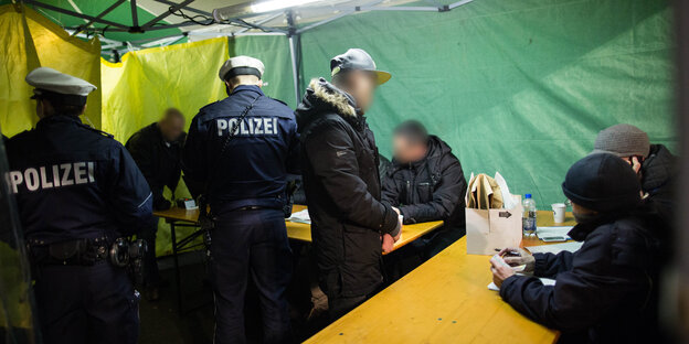 Polizisten kontrollieren Verdächtige an Tischen in einem grünen Zelt Polizisten kontrollieren Verdächtige an Tischen in einem grünen Zelt