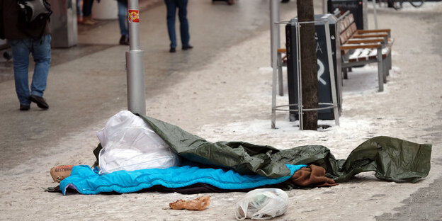 Auf einem Gehweg liegt ein Schlafsack im Schnee, dahinter eine Parkbank, Menschen laufen vorüber Auf einem Gehweg liegt ein Schlafsack im Schnee, dahinter eine Parkbank, Menschen laufen vorüber
