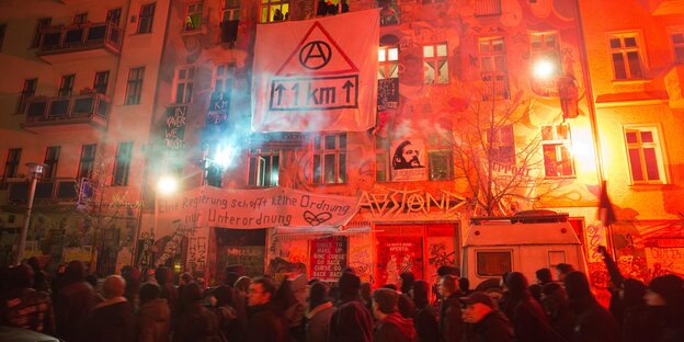 Dunkel gekleidete Menschen demonstriren auf der Straße. Die Häuser dahinter leuchten rot-orange und an den Häusern hängen Plakate. Dunkel gekleidete Menschen demonstriren auf der Straße. Die Häuser dahinter leuchten rot-orange und an den Häusern hängen Plakate.